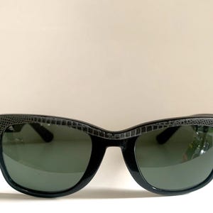 May include: Black sunglasses with a textured gray pattern on the top frame and dark green lenses. The sunglasses have a classic, retro design, suitable for eye protection from the sun.
