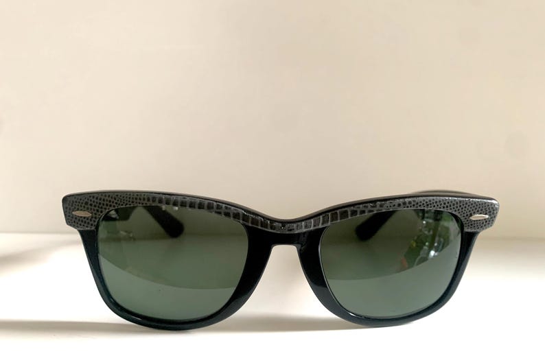 May include: Black sunglasses with a textured gray pattern on the top frame and dark green lenses. The sunglasses have a classic, retro design, suitable for eye protection from the sun.