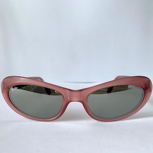 Peut inclure: Lunettes de soleil &agrave; monture rose avec verres gris. Les lunettes de soleil ont une forme incurv&eacute;e en &oelig;il de chat. La monture est de couleur rose translucide. Les verres sont de couleur gris fonc&eacute;. Les lunettes sont pos&eacute;es sur un fond blanc.