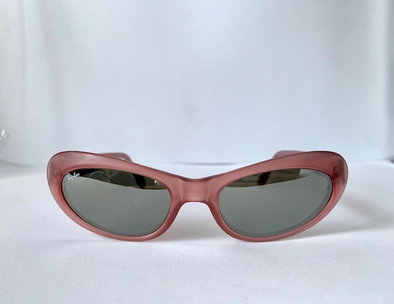 Peut inclure: Lunettes de soleil &agrave; monture rose avec verres gris. Les lunettes de soleil ont une forme incurv&eacute;e en &oelig;il de chat. La monture est de couleur rose translucide. Les verres sont de couleur gris fonc&eacute;. Les lunettes sont pos&eacute;es sur un fond blanc.
