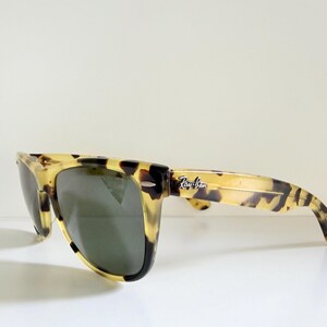 May include: Tortoiseshell-patterned sunglasses with dark lenses. The sunglasses have a classic rectangular frame and the Ray-Ban logo is visible on the temple. The glasses are set against a white background.