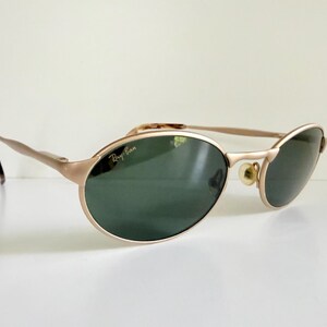 Puede incluir: Gafas de sol Ray-Ban doradas con lentes ovaladas y tinte verde oscuro. Las monturas tienen un brillo met&aacute;lico sutil y las patillas presentan un patr&oacute;n de carey. El logotipo de Ray-Ban es visible en la lente.