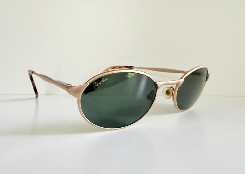 Puede incluir: Gafas de sol Ray-Ban doradas con lentes ovaladas y tinte verde oscuro. Las monturas tienen un brillo met&aacute;lico sutil y las patillas presentan un patr&oacute;n de carey. El logotipo de Ray-Ban es visible en la lente.