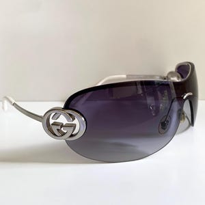 May include: Gucci sunglasses featuring a rimless, wrap-around dark gradient lens. The silver-tone metal temples are accented with a prominent interlocking G logo at the hinge, transitioning into white temple tips for a sleek, modern eyewear design.