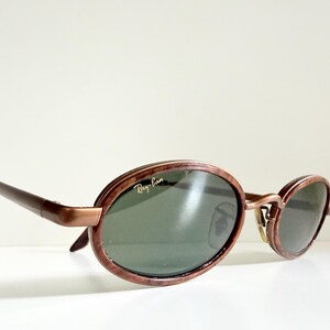 May include: Oval sunglasses with a brown frame and dark green lenses. The Ray-Ban logo is visible on the lens. The sunglasses have a retro style.