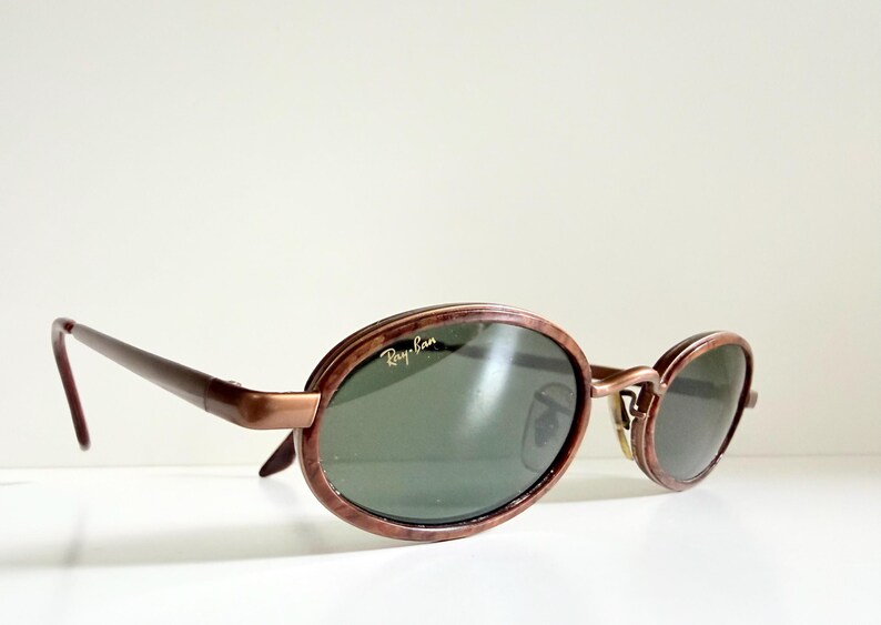 May include: Oval sunglasses with a brown frame and dark green lenses. The Ray-Ban logo is visible on the lens. The sunglasses have a retro style.