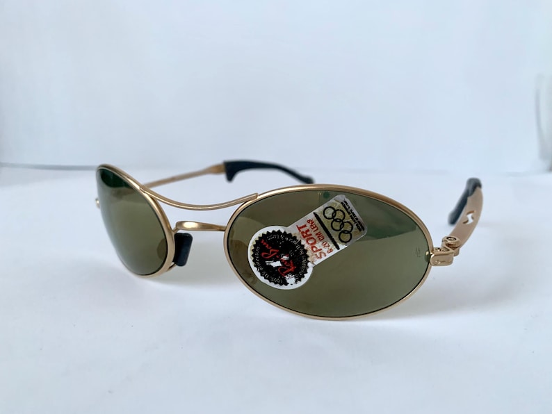 May include: Gold-tone sunglasses with oval lenses and black accents. The sunglasses have a sporty design and a sticker with the text "SPORT" and the Olympic rings.