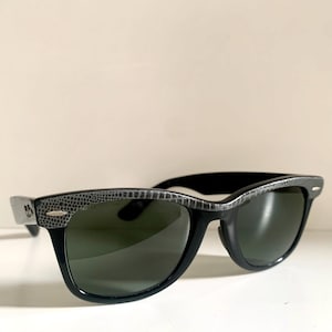 May include: Black sunglasses with dark green lenses. The frames are black with a textured, gray pattern along the top and sides. The sunglasses are set against a white background.