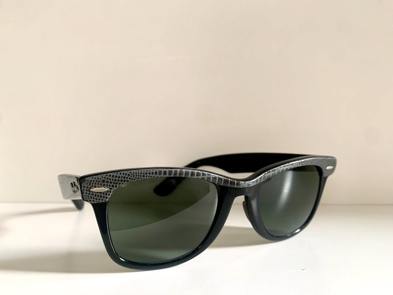 May include: Black sunglasses with dark green lenses. The frames are black with a textured, gray pattern along the top and sides. The sunglasses are set against a white background.
