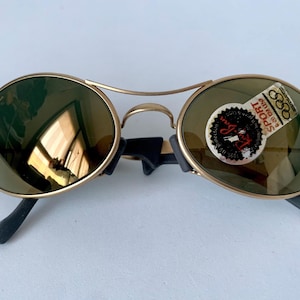 May include: Gold-framed sunglasses with round lenses and black temples. The lenses have a green tint, and a sticker with text and Olympic rings is on one lens. The sunglasses are on a white surface.