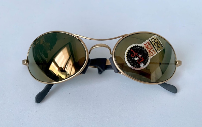May include: Gold-framed sunglasses with round lenses and black temples. The lenses have a green tint, and a sticker with text and Olympic rings is on one lens. The sunglasses are on a white surface.