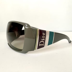 Peut inclure: Lunettes de soleil grises avec des verres foncés. Les lunettes ont une monture épaisse avec une bande bordeaux, sarcelle et blanche. Le mot "Dior" est imprimé sur le côté de la monture.