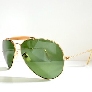 May include: Gold-framed aviator sunglasses with green lenses. The sunglasses have a double bridge and a light brown bar across the top. The temples are thin and gold-colored. The lenses are a teardrop shape.