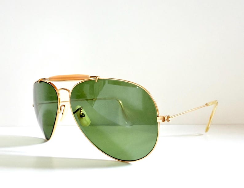 May include: Gold-framed aviator sunglasses with green lenses. The sunglasses have a double bridge and a light brown bar across the top. The temples are thin and gold-colored. The lenses are a teardrop shape.
