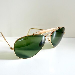 May include: Aviator-style sunglasses with a gold-colored frame and green lenses. The sunglasses have a double bridge and a curved brow bar. The Ray-Ban logo is visible on the lens. The glasses are set against a white background.
