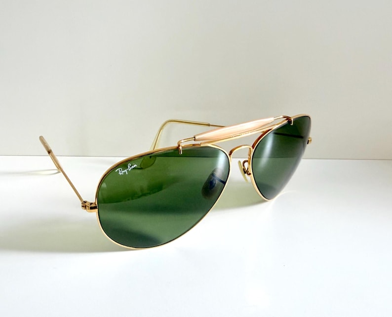 May include: Aviator-style sunglasses with a gold-colored frame and green lenses. The sunglasses have a double bridge and a curved brow bar. The Ray-Ban logo is visible on the lens. The glasses are set against a white background.