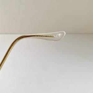 May include: Close-up of a gold-toned metal eyeglass frame arm with a clear plastic earpiece. The text "BOUDOIR" is printed on the earpiece. The arm curves gently, and the background is a plain white.