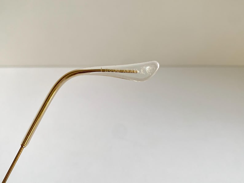 May include: Close-up of a gold-toned metal eyeglass frame arm with a clear plastic earpiece. The text "BOUDOIR" is printed on the earpiece. The arm curves gently, and the background is a plain white.