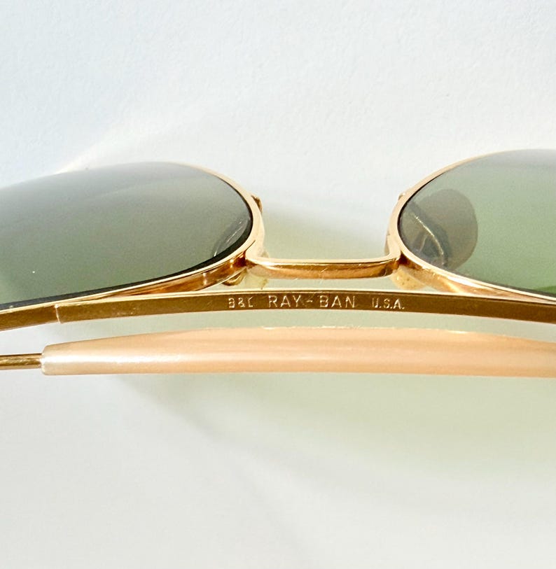 May include: Close-up of Ray-Ban sunglasses with gold frames and green lenses. The text "B&L RAY-BAN U.S.A." is printed on the frame. The sunglasses have a classic aviator style.