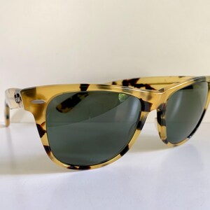May include: Tortoiseshell sunglasses with dark green lenses. The frames have a classic, rectangular shape with a speckled pattern in shades of tan and brown. The sunglasses are angled slightly, showcasing the design and style.