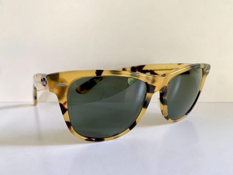 May include: Tortoiseshell sunglasses with dark green lenses. The frames have a classic, rectangular shape with a speckled pattern in shades of tan and brown. The sunglasses are angled slightly, showcasing the design and style.