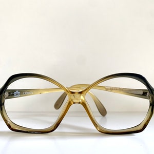 Könnte beinhalten: Eine Brille in Braun- und Goldtönen mit einem geometrischen Rahmen. Die Brille hat einen Farbverlauf, der von Dunkelbraun zu Hellgold übergeht. Der Markenname "COBRA" ist auf dem linken Bügel aufgedruckt.