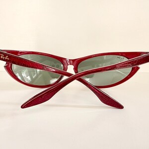 May include: Red-framed sunglasses with a glittery finish. The sunglasses have a cat-eye shape and gray lenses. The Ray-Ban logo is visible on the left arm. The arms are crossed, showing the back of the sunglasses.