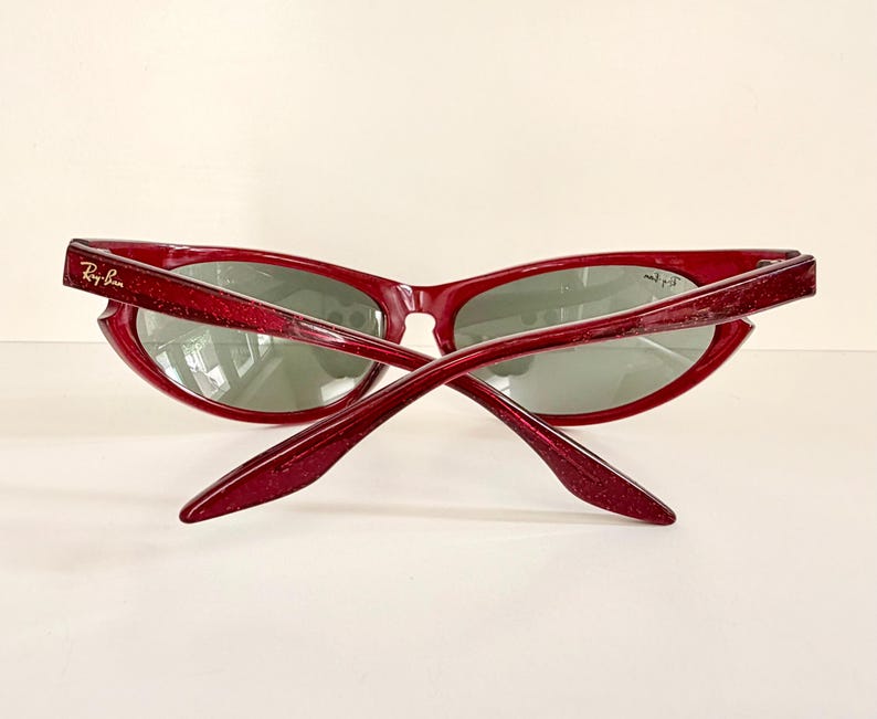May include: Red-framed sunglasses with a glittery finish. The sunglasses have a cat-eye shape and gray lenses. The Ray-Ban logo is visible on the left arm. The arms are crossed, showing the back of the sunglasses.