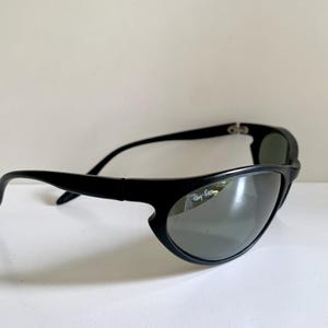 May include: Black sunglasses with a wrap-around frame design and dark tinted lenses. The right lens features the white Ray-Ban logo printed in the upper corner. The temples are curved and solid black, connecting to a sturdy, continuous frame front.
