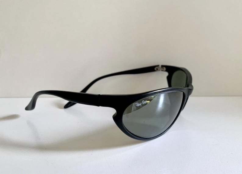 May include: Black sunglasses with a wrap-around frame design and dark tinted lenses. The right lens features the white Ray-Ban logo printed in the upper corner. The temples are curved and solid black, connecting to a sturdy, continuous frame front.
