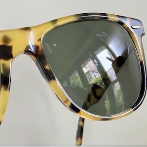 May include: Tortoiseshell sunglasses with dark green lenses. The frames have a classic design with a speckled pattern in shades of brown and yellow. The lenses reflect the surrounding environment.
