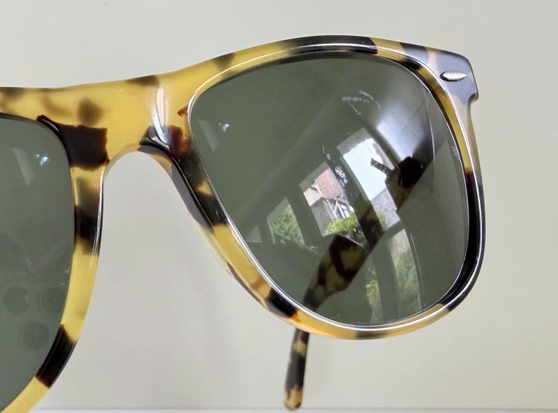 May include: Tortoiseshell sunglasses with dark green lenses. The frames have a classic design with a speckled pattern in shades of brown and yellow. The lenses reflect the surrounding environment.