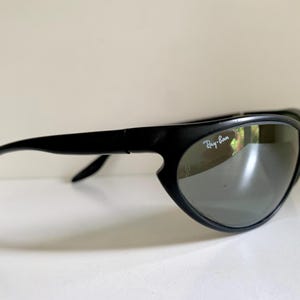 May include: Black sunglasses with a matte finish and a wraparound frame design. The lenses are dark grey and feature the white Ray-Ban logo printed on the upper right corner. The temples are curved and extend from the outer edges of the frames.