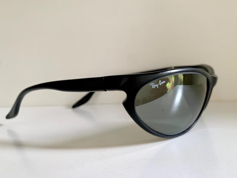 May include: Black sunglasses with a matte finish and a wraparound frame design. The lenses are dark grey and feature the white Ray-Ban logo printed on the upper right corner. The temples are curved and extend from the outer edges of the frames.