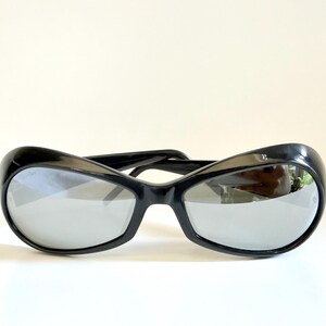 May include: Black sunglasses with a curved, oversized frame and reflective lenses. The sunglasses have a sleek, modern design, suitable for eye protection from the sun. The lenses are a mirrored silver colour.