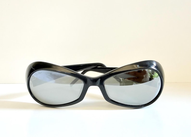 May include: Black sunglasses with a curved, oversized frame and reflective lenses. The sunglasses have a sleek, modern design, suitable for eye protection from the sun. The lenses are a mirrored silver colour.