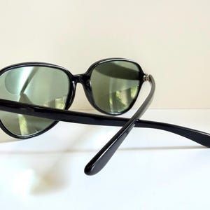 May include: Black sunglasses with oval frames and green lenses. The sunglasses have a thick, glossy black frame and temples. The arms of the sunglasses are extended, showing the design of the temples.