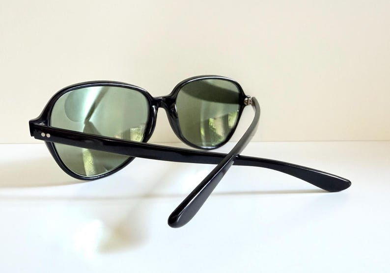 May include: Black sunglasses with oval frames and green lenses. The sunglasses have a thick, glossy black frame and temples. The arms of the sunglasses are extended, showing the design of the temples.