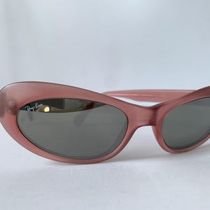 Peut inclure: Lunettes de soleil roses avec des verres gris fonc&eacute;. Les lunettes de soleil ont une forme arrondie en &oelig;il de chat et le logo Ray-Ban est visible sur la lentille gauche. Les montures sont de couleur rose translucide unie.