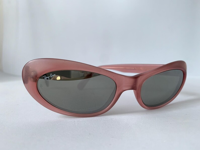 Peut inclure: Lunettes de soleil roses avec des verres gris fonc&eacute;. Les lunettes de soleil ont une forme arrondie en &oelig;il de chat et le logo Ray-Ban est visible sur la lentille gauche. Les montures sont de couleur rose translucide unie.