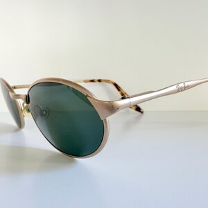 Puede incluir: Gafas de sol Ray-Ban doradas con lentes verdes. Las monturas tienen una forma de aviador cl&aacute;sica con una curva sutil. Las patillas son de color dorado claro con el logotipo de Ray-Ban y puntas carey.