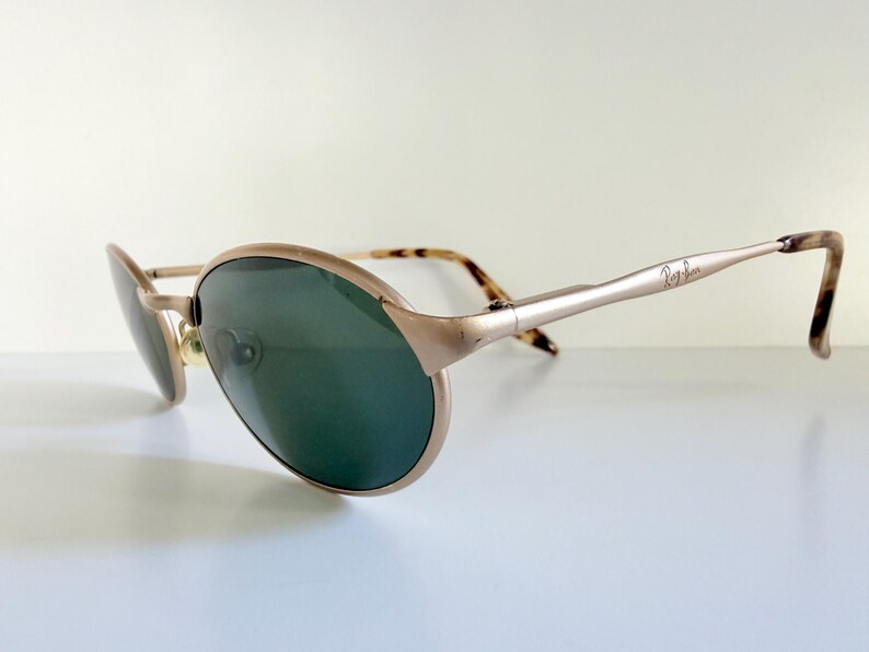 Puede incluir: Gafas de sol Ray-Ban doradas con lentes verdes. Las monturas tienen una forma de aviador cl&aacute;sica con una curva sutil. Las patillas son de color dorado claro con el logotipo de Ray-Ban y puntas carey.