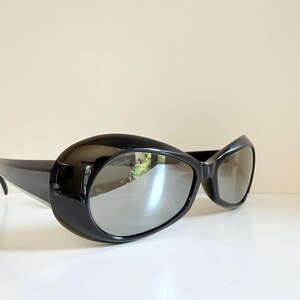 May include: Black sunglasses with a rounded, oversized frame and reflective lenses. The sunglasses have a sleek, modern design, with a curved shape and thick temples. The lenses reflect the surrounding environment.