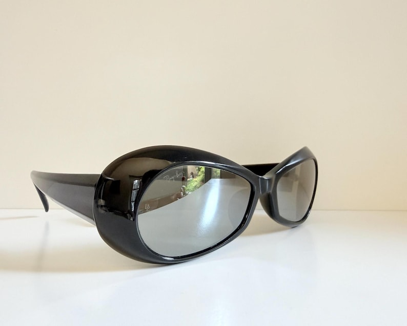May include: Black sunglasses with a rounded, oversized frame and reflective lenses. The sunglasses have a sleek, modern design, with a curved shape and thick temples. The lenses reflect the surrounding environment.
