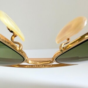 May include: Close-up of gold-rimmed sunglasses with green lenses. The sunglasses feature light-colored nose pads and a gold bridge. The image highlights the intricate details of the frame and the lenses.