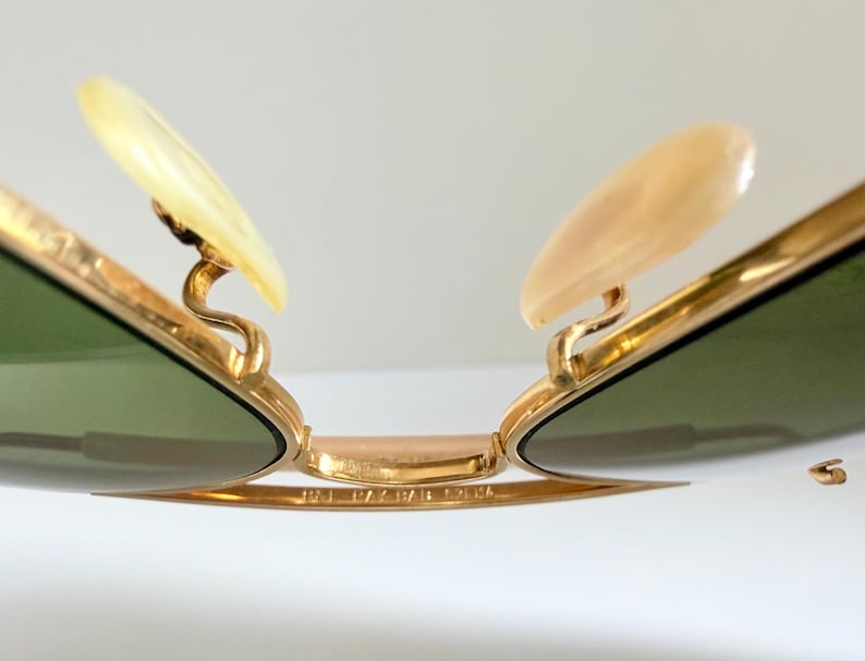 May include: Close-up of gold-rimmed sunglasses with green lenses. The sunglasses feature light-colored nose pads and a gold bridge. The image highlights the intricate details of the frame and the lenses.