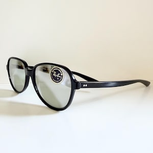 May include: Black sunglasses with reflective lenses. The sunglasses have a thick, rounded frame and straight arms. A circular sticker with the words "Ray-Ban" is on the left lens. The sunglasses are on a white surface.