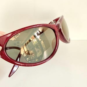 May include: Burgundy-colored sunglasses with a speckled finish. The sunglasses have a rounded frame and dark lenses. The Ray-Ban logo is visible on the left lens. The sunglasses are angled, showing the side of the frame and the reflection of a room.