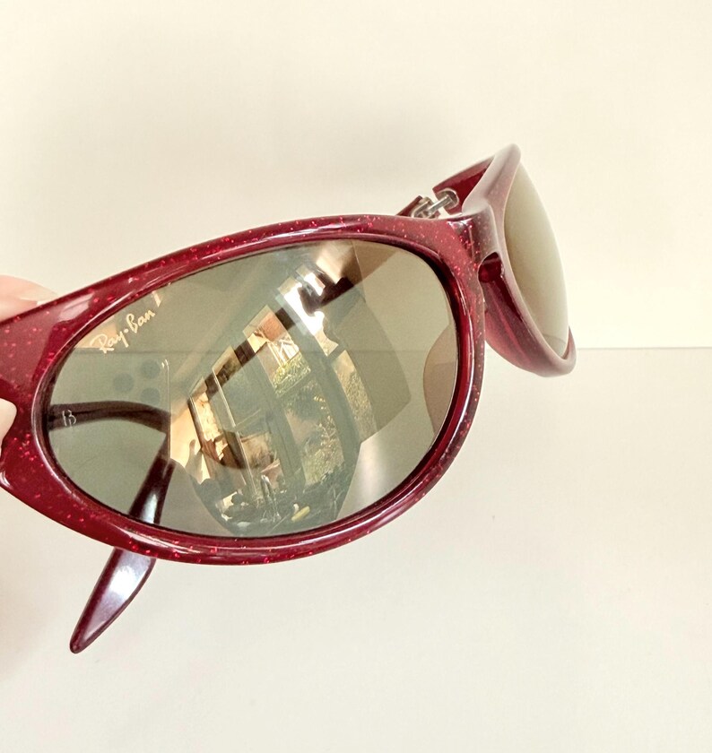 May include: Burgundy-colored sunglasses with a speckled finish. The sunglasses have a rounded frame and dark lenses. The Ray-Ban logo is visible on the left lens. The sunglasses are angled, showing the side of the frame and the reflection of a room.