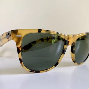 May include: Tortoiseshell sunglasses with dark green lenses. The frames have a classic design with a speckled pattern in shades of brown and yellow. The sunglasses are set against a white background.